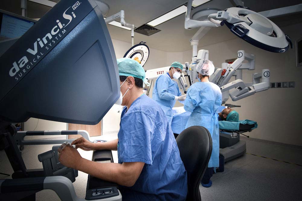 Tres médicos dentro de un quirófano en un procedimiento de cirugía robótica. Uno sentado frente a la consola de control y dos operando.