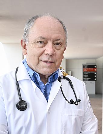 Es la foto de perfil de un pediatra en León de Hospital Angeles León. Usa bata blanca con camisa azul, tiene un estetoscopio en los hombros y de fondo se ve el pasillo del hospital.